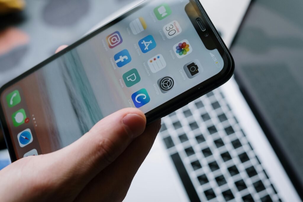 Person holding a smartphone in front of a laptop showcasing various app icons, highlighting digital connectivity.